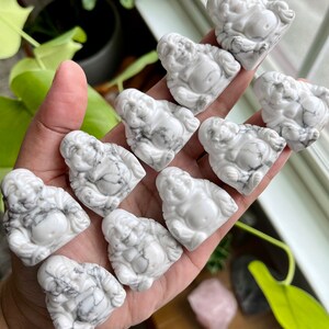 May include: A collection of ten white and gray carved stone figurines of a laughing Buddha. The figurines are arranged in a hand, with the largest figurine at the top and the smallest figurine at the bottom.