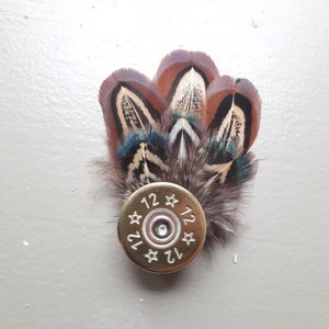 Peut inclure: Broche artisanale composée de plumes marron, bleues et blanches, fixées sur une douille de cartouche de fusil de calibre 12 dorée. Le numéro "12" est estampillé sur le bord de la douille.