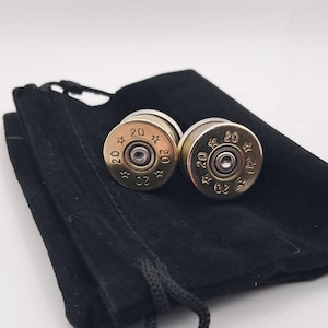 May include: A pair of brass-coloured cufflinks, each bearing the number "20" and star motifs, rest on a black velvet pouch. The cufflinks have a metallic sheen and a detailed, industrial aesthetic.