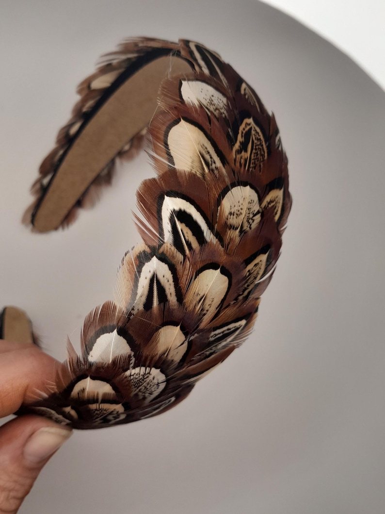 May include: A decorative headband featuring a brown and white feather design. The feathers are arranged in a layered pattern, creating a textured and eye-catching accessory. The headband is made of a sturdy material and is suitable for special occasions or everyday wear.