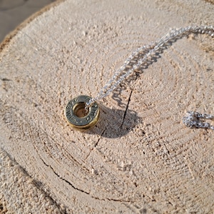 May include: A gold-coloured pendant necklace with a silver chain. The pendant is a small, circular ring with engraved text. The necklace is displayed on a light brown, textured wooden surface.
