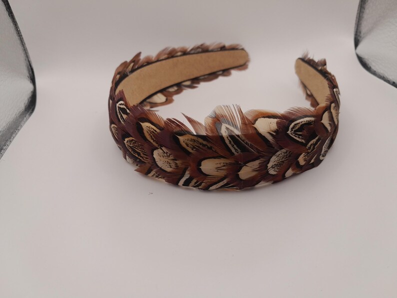 May include: A brown and cream-colored feather headband. The feathers are arranged around a brown band. The headband is a hair accessory, likely for special occasions or costume use. The feathers have a natural, textured appearance.