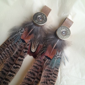 Puede incluir: Pendientes hechos a mano con plumas de faisán en tonos marrón, negro y turquesa. Las plumas están unidas a una correa de cuero y a una vaina de bala plateada. Un accesorio único para un look rústico.