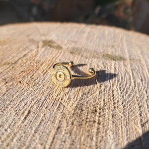 May include: A gold-coloured ring with a repurposed bullet casing as its focal point. The casing is engraved with the numbers "5.56" and "GGG". The ring has a simple, adjustable band design.