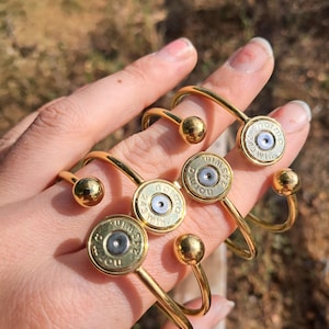 Puede incluir: Tres pulseras abiertas de tono dorado, cada una con un culote de casquillo de latón como pieza central. Los culotes están grabados con el texto "Norma" y "243 Win" alrededor de un fulminante central plateado. Cada pulsera termina en una pequeña bola dorada.