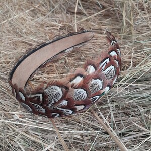 May include: A decorative headband featuring brown and white patterned feathers. The headband has a tan suede interior and is surrounded by a band of feathers. The feathers have a mix of brown, white, and black markings.