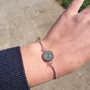 Könnte beinhalten: Ein silbernes Armband mit einer wiederverwendeten Patronenhülse als zentralem Anhänger. Die Hülse hat eine metallische Oberfläche mit sichtbaren Markierungen. Das Armband ist filigran und hat ein schlichtes Ketten-Design.
