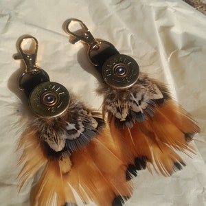 May include: Two keychains with gold-colored clasps, black leather straps, and brass-colored bullet casings. Each keychain features a cluster of brown, tan, and black feathers, creating a rustic and unique accessory.
