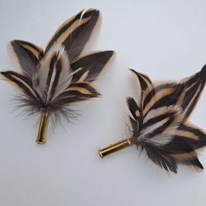 Puede incluir: Dos boutonnieres decorativas de plumas. Cada boutonniere presenta un grupo de plumas marrones, beige y negras unidas a una vaina de bala dorada. Las plumas están dispuestas en forma floral estilizada.