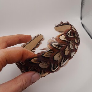 May include: A brown and white feather headband. The headband is made of feathers in shades of brown, tan, and white. The feathers are attached to a brown band. The headband is held by a hand.