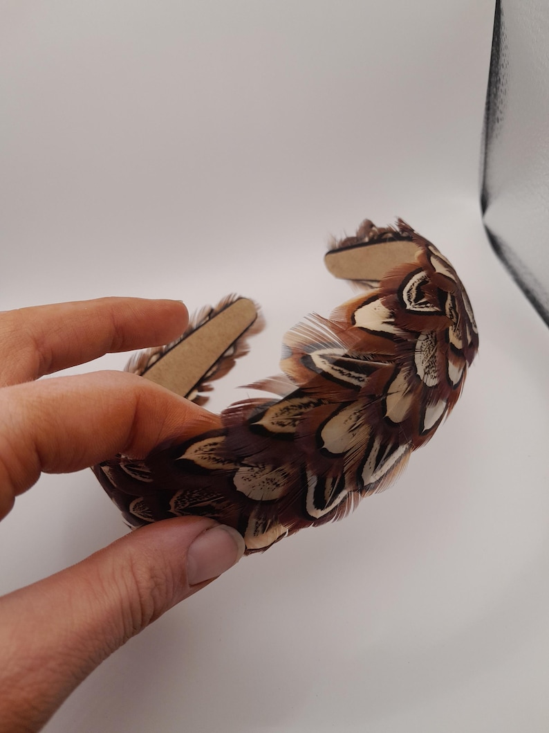 May include: A brown and white feather headband. The headband is made of feathers in shades of brown, tan, and white. The feathers are attached to a brown band. The headband is held by a hand.