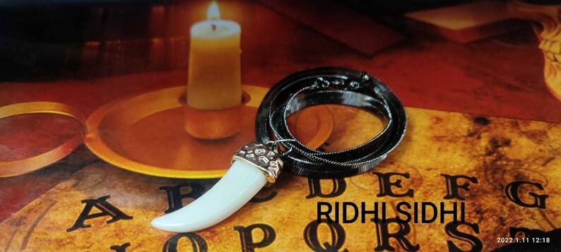 May include: A white fang pendant necklace with a silver chain. The pendant is on a black and gold Ouija board with the letters A, B, C, D, E, F, G, R, I, D, H, L, S, I, D, H, L, Q, R, S.