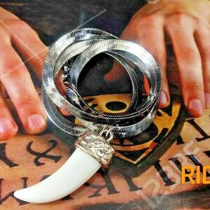 May include: A silver snake chain necklace with a white tooth pendant. The necklace is lying on a surface with letters printed on it. The text on the surface reads "RIDHI SIDHI".