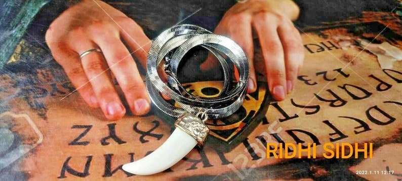 May include: A silver snake chain necklace with a white tooth pendant. The necklace is lying on a surface with letters printed on it. The text on the surface reads "RIDHI SIDHI".