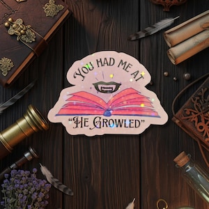 May include: A sticker with the text "You had me at HE GROWLED" over an open book with a vampire mouth. The sticker is surrounded by vintage books, feathers, and a telescope on a dark wooden surface.