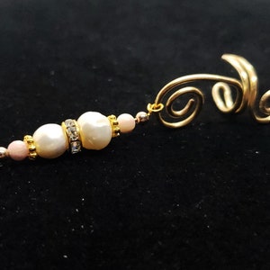 May include: Gold-toned ear cuff with a dangling beaded accent. The accent features two large, off-white pearls, smaller pink beads, and a band of clear rhinestones. The cuff has a swirling design.
