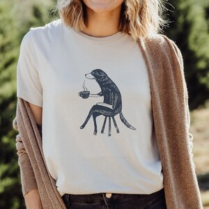 Puede incluir: Una mujer con una camiseta blanca con un gr&aacute;fico de un perro sosteniendo una taza de caf&eacute;. El perro est&aacute; sentado en una silla. La mujer lleva un c&aacute;rdigan marr&oacute;n y pantalones vaqueros negros. Est&aacute; de pie en un campo de &aacute;rboles.
