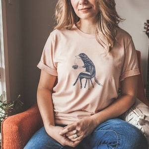 Puede incluir: Una mujer lleva una camiseta rosa claro con un gr&aacute;fico de un perro sentado en una silla y sosteniendo una taza de caf&eacute;. El perro es blanco y negro y el gr&aacute;fico es de estilo vintage.