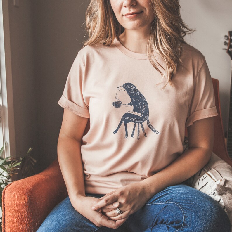 Puede incluir: Una mujer lleva una camiseta rosa claro con un gr&aacute;fico de un perro sentado en una silla y sosteniendo una taza de caf&eacute;. El perro es blanco y negro y el gr&aacute;fico es de estilo vintage.