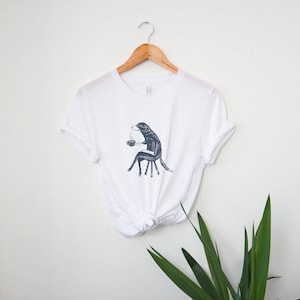 Puede incluir: Camiseta blanca con una ilustraci&oacute;n en blanco y negro de un perro sentado en una silla y sosteniendo una taza de caf&eacute;.