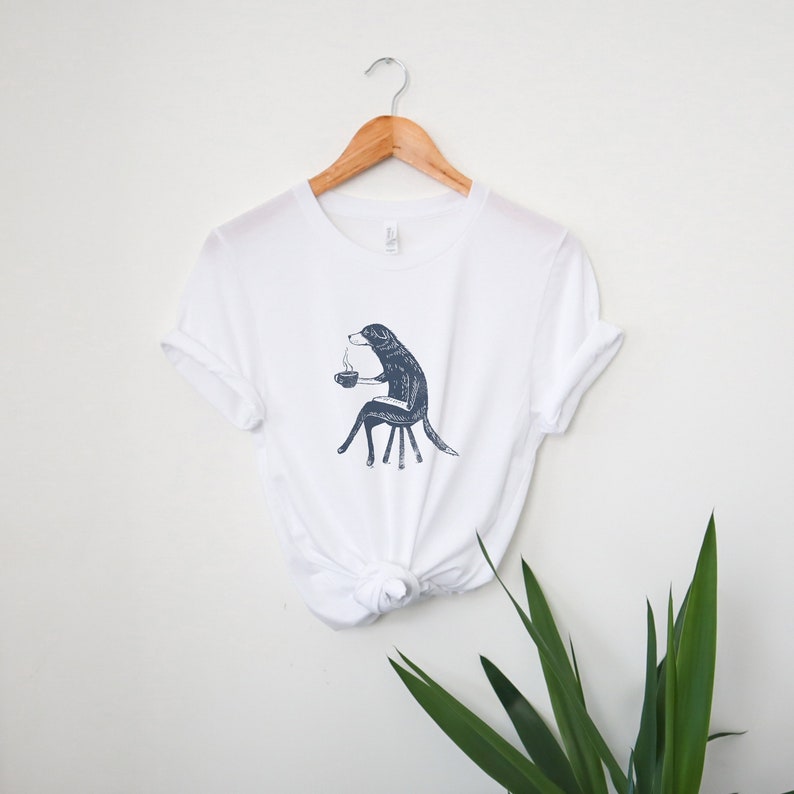 Puede incluir: Camiseta blanca con una ilustraci&oacute;n en blanco y negro de un perro sentado en una silla y sosteniendo una taza de caf&eacute;.