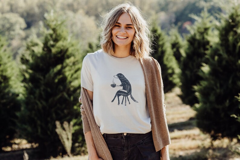 Puede incluir: Una mujer con una camiseta blanca con un gr&aacute;fico de un perro sosteniendo una taza de caf&eacute;. El perro est&aacute; sentado en una silla. La mujer lleva un c&aacute;rdigan marr&oacute;n y pantalones vaqueros negros. Est&aacute; de pie en un campo de &aacute;rboles.