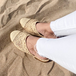 以下が含まれることがあります： 丸いつま先のベージュ色の織りスリッポンシューズのペア。靴は明るい色のオープン織りの素材で作られています。靴は白いズボンと合わせて着用されており、背景は砂です。