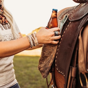 Op de afbeelding: Een bruine bierfles met een blauwe dop wordt vastgehouden in de buurt van een bruin leren zadel. Het zadel heeft decoratieve stiksels en metalen hardware. Een persoon met een lichtgekleurd shirt en armbanden houdt de fles vast.