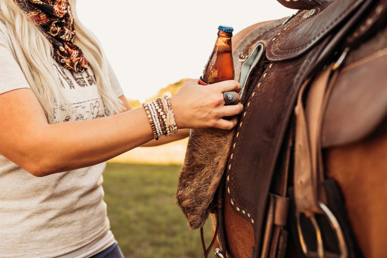Op de afbeelding: Een bruine bierfles met een blauwe dop wordt vastgehouden in de buurt van een bruin leren zadel. Het zadel heeft decoratieve stiksels en metalen hardware. Een persoon met een lichtgekleurd shirt en armbanden houdt de fles vast.