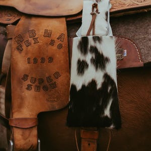 Op de afbeelding: Een close-up van een bruin leren zadel met een koeienhuid accessoire. Het zadel heeft reli&euml;fdetails en de tekst "N B H A TEX 20 1999 SUPER SHOW". Het koeienhuid accessoire is zwart en wit.