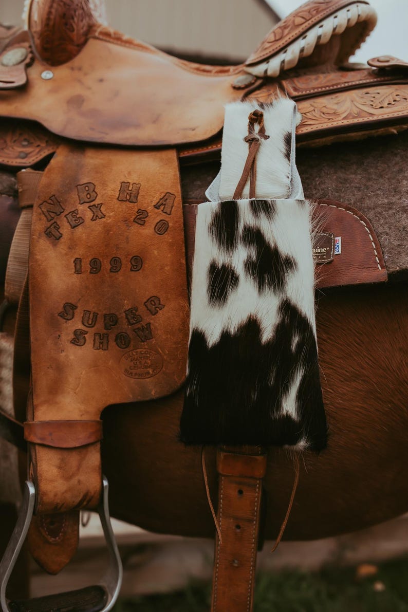 Op de afbeelding: Een close-up van een bruin leren zadel met een koeienhuid accessoire. Het zadel heeft reli&euml;fdetails en de tekst "N B H A TEX 20 1999 SUPER SHOW". Het koeienhuid accessoire is zwart en wit.
