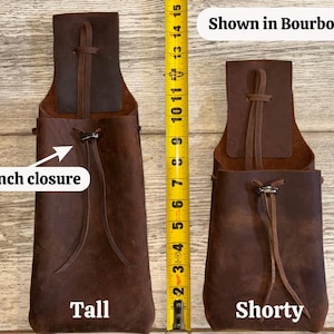 Op de afbeelding: Twee bruine leren zakjes, een lange en een korte, met trekkoordsluitingen. De lange zak meet ongeveer 35,5 cm in hoogte. De tekst "Shown in Bourbon" wordt boven het kortere zakje weergegeven. Een gele rolmaat is naast de zakjes geplaatst.