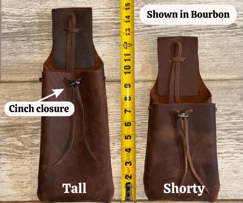 Op de afbeelding: Twee bruine leren zakjes, een lange en een korte, met trekkoordsluitingen. De lange zak meet ongeveer 35,5 cm in hoogte. De tekst "Shown in Bourbon" wordt boven het kortere zakje weergegeven. Een gele rolmaat is naast de zakjes geplaatst.