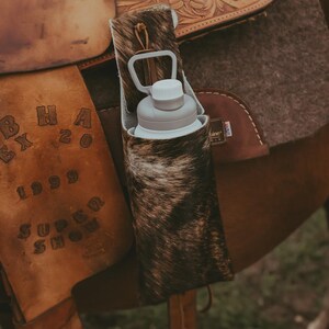 Op de afbeelding: Een waterfleshouder van koeienhuid met een grijze waterfles erin, bevestigd aan een bruin leren zadel. Het zadel heeft ingewikkeld gereedschap en staat op een paard. De houder is gemaakt van bruine en witte koeienhuid.