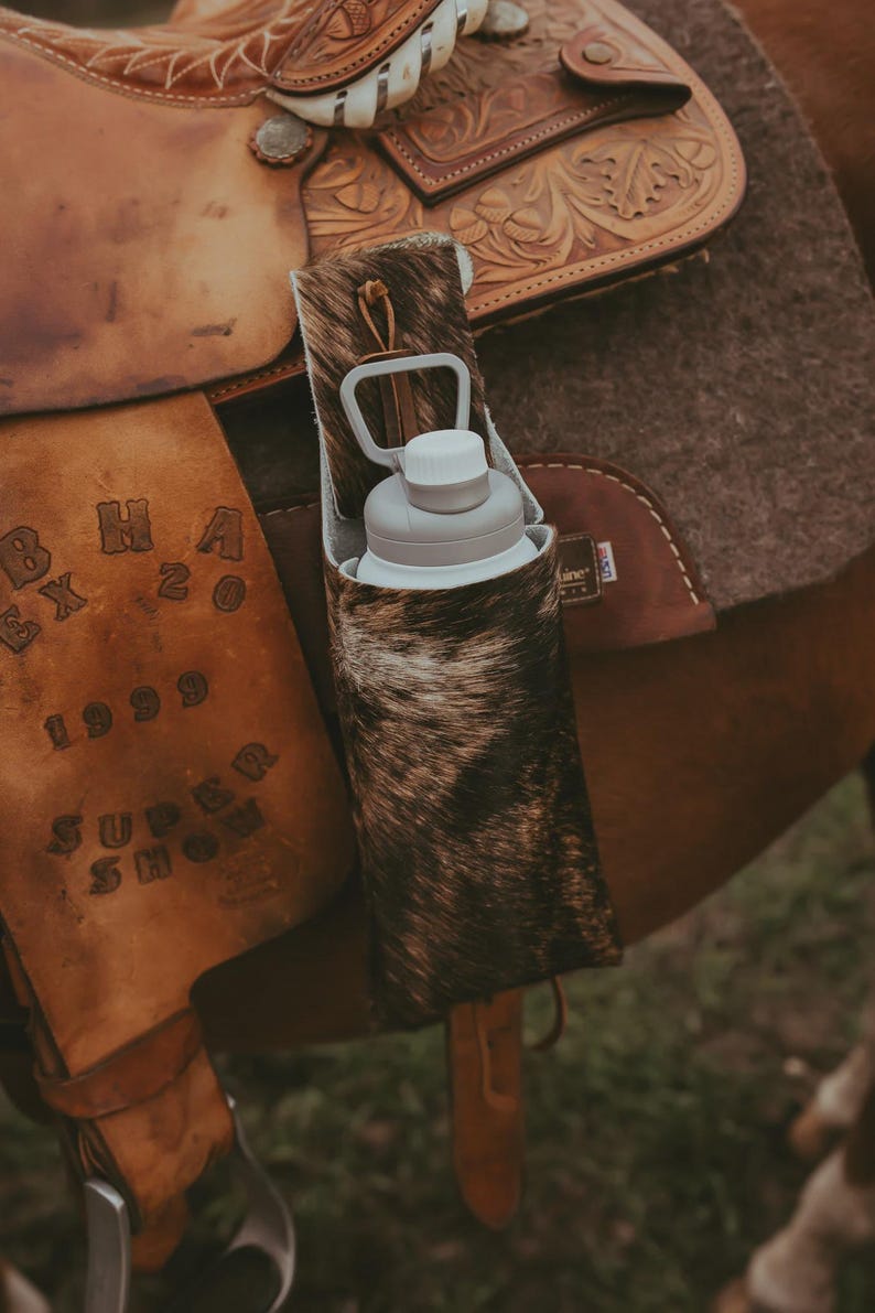 Op de afbeelding: Een waterfleshouder van koeienhuid met een grijze waterfles erin, bevestigd aan een bruin leren zadel. Het zadel heeft ingewikkeld gereedschap en staat op een paard. De houder is gemaakt van bruine en witte koeienhuid.