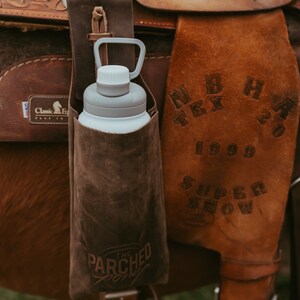 Op de afbeelding: Een bruine leren waterfleshouder met een witte en grijze waterfles bevestigd aan een bruin leren zadel. De houder heeft de tekst "The Parched Pony" erop. Het zadel heeft de tekst "N B H TEX 20 1999 SUPER SHOW".