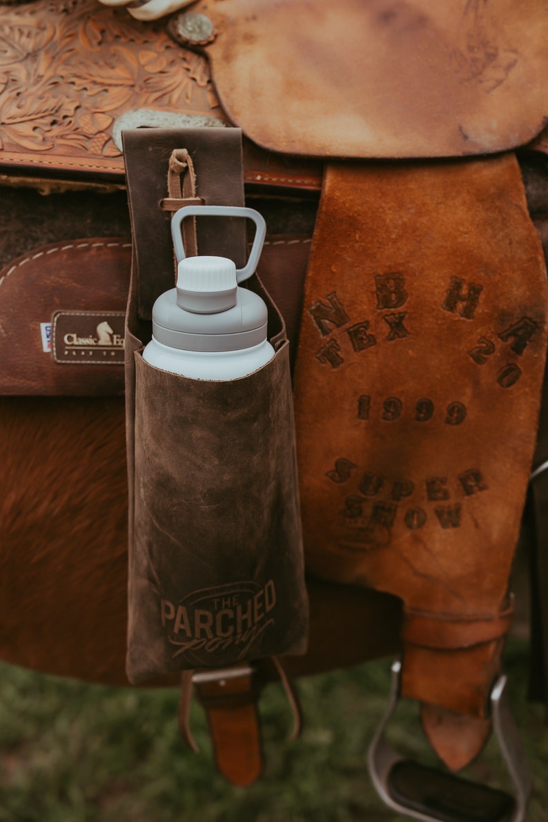 Op de afbeelding: Een bruine leren waterfleshouder met een witte en grijze waterfles bevestigd aan een bruin leren zadel. De houder heeft de tekst "The Parched Pony" erop. Het zadel heeft de tekst "N B H TEX 20 1999 SUPER SHOW".