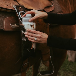 May include: A white and black cowhide water bottle holder attached to a brown leather saddle on a brown horse. The holder has a white water bottle inside.