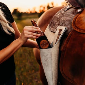 Op de afbeelding: Een bruine glazen fles met een zwarte koozie met de tekst "PARCHED" wordt vastgehouden in de buurt van een paardenzadel. Het zadel is bruin leer met decoratieve bewerkingen. Er hangt een lichtgekleurde zadeltas.
