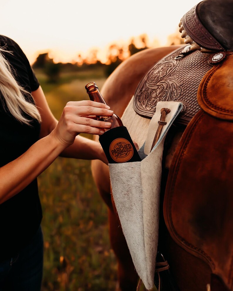 Op de afbeelding: Een bruine glazen fles met een zwarte koozie met de tekst "PARCHED" wordt vastgehouden in de buurt van een paardenzadel. Het zadel is bruin leer met decoratieve bewerkingen. Er hangt een lichtgekleurde zadeltas.