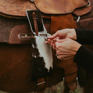 Op de afbeelding: Een close-up van een bruin leren zadel met ingewikkelde details en een koeienhuid accessoire. Het zadel staat op een paard, met handen die een kleine leren riem vasthouden. De sc&egrave;ne speelt zich buiten af.