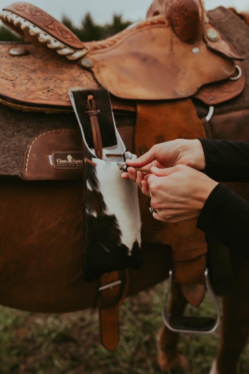 Op de afbeelding: Een close-up van een bruin leren zadel met ingewikkelde details en een koeienhuid accessoire. Het zadel staat op een paard, met handen die een kleine leren riem vasthouden. De sc&egrave;ne speelt zich buiten af.