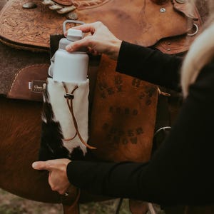 Op de afbeelding: Een witte waterfles met een grijze dop wordt vastgehouden in een koeienhuidhouder die aan een bruin leren zadel is bevestigd. De houder heeft een leren trekkoord. Het zadel staat op een bruin paard.