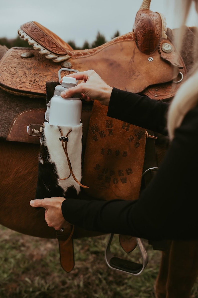 Op de afbeelding: Een witte waterfles met een grijze dop wordt vastgehouden in een koeienhuidhouder die aan een bruin leren zadel is bevestigd. De houder heeft een leren trekkoord. Het zadel staat op een bruin paard.