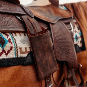 Op de afbeelding: Detailopname van een bruin leren zadel met een bijpassend leren etui. Het zadel heeft ingewikkelde details en is versierd met een kleurrijk patroon. Het etui, vastgemaakt met een leren veter, toont een rustieke uitstraling.