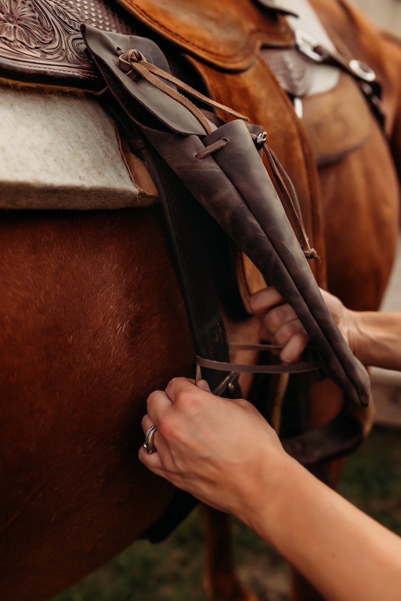 Op de afbeelding: Close-up van een bruin leren zadel op een paard. Het zadel heeft ingewikkelde details en wordt vastgezet met riemen. Een donkerbruine leren schede is aan het zadel bevestigd. De vacht van het paard heeft een rijke, roodbruine kleur.