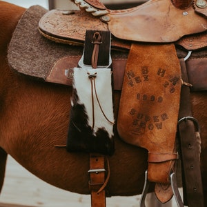 May include: A brown leather saddle with intricate tooling and a cowhide pouch with black and white markings. The saddle is on a brown horse, with a leather stirrup and a leather strap.