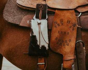 Può includere: Una sella in pelle marrone con lavorazione intricata e una custodia in pelle di mucca con macchie bianche e nere. La sella è su un cavallo marrone, con una staffa in pelle e un cinturino in pelle.