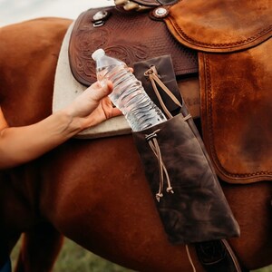 Op de afbeelding: Een bruine leren waterfleshouder bevestigd aan een paardenzadel. De houder is ontworpen om een standaard waterfles te dragen. Het zadel is bruin leer met decoratieve bewerkingen. Een doorzichtige plastic waterfles wordt in de houder gehouden.