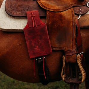 Op de afbeelding: Close-up van een bruin leren zadel met een rode leren tasje bevestigd. Het zadel staat op een bruin paard. De stijgbeugel is zichtbaar. Het zadel heeft een witte pad.